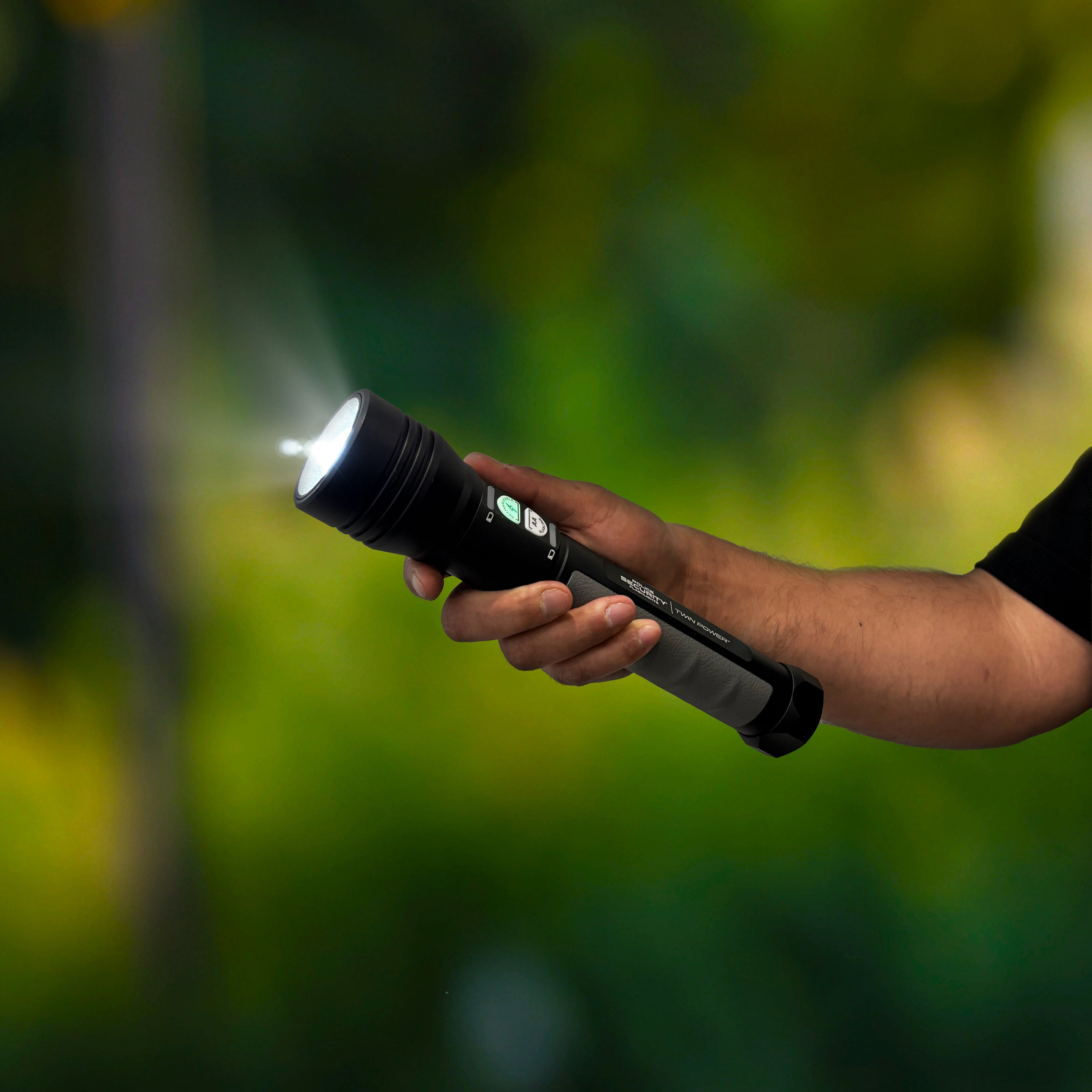 A hand holding a Police Security Twin Power flashlight that is turned on, emitting a bright beam of light, set against a blurred green outdoor background.