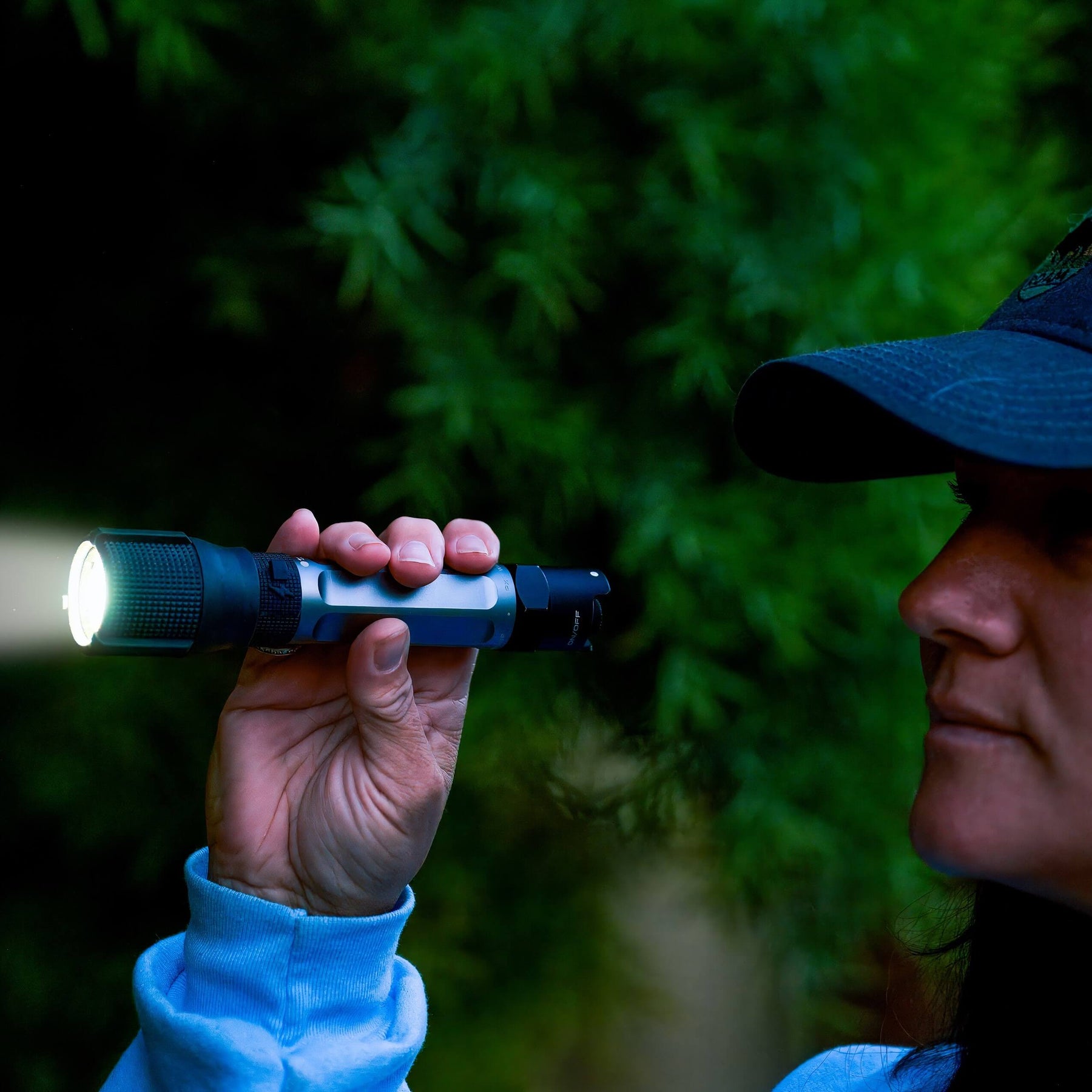 A person in a blue cap and light blue hoodie holds a black Police Security Zephyr-R tactical flashlight with its bright LED turned on, pointing it to the left, against a dark, blurry background of green foliage.