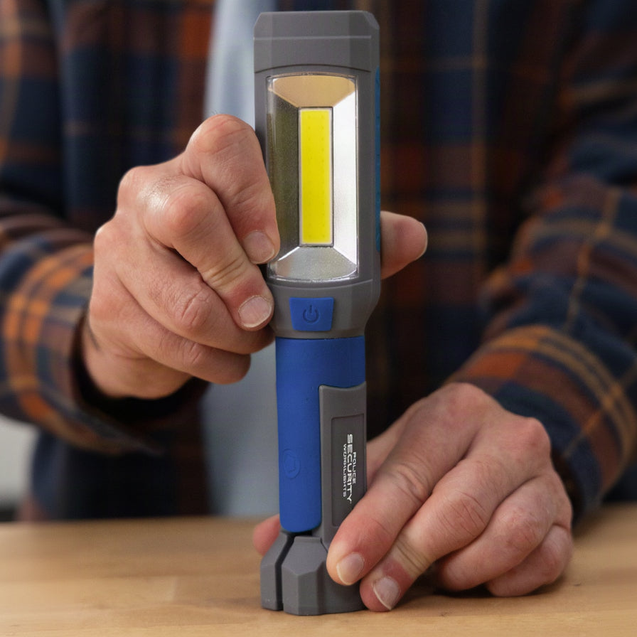 A person holding a compact LED work light with a gray and blue body, featuring a large yellow LED chip on the front, over a wooden surface.