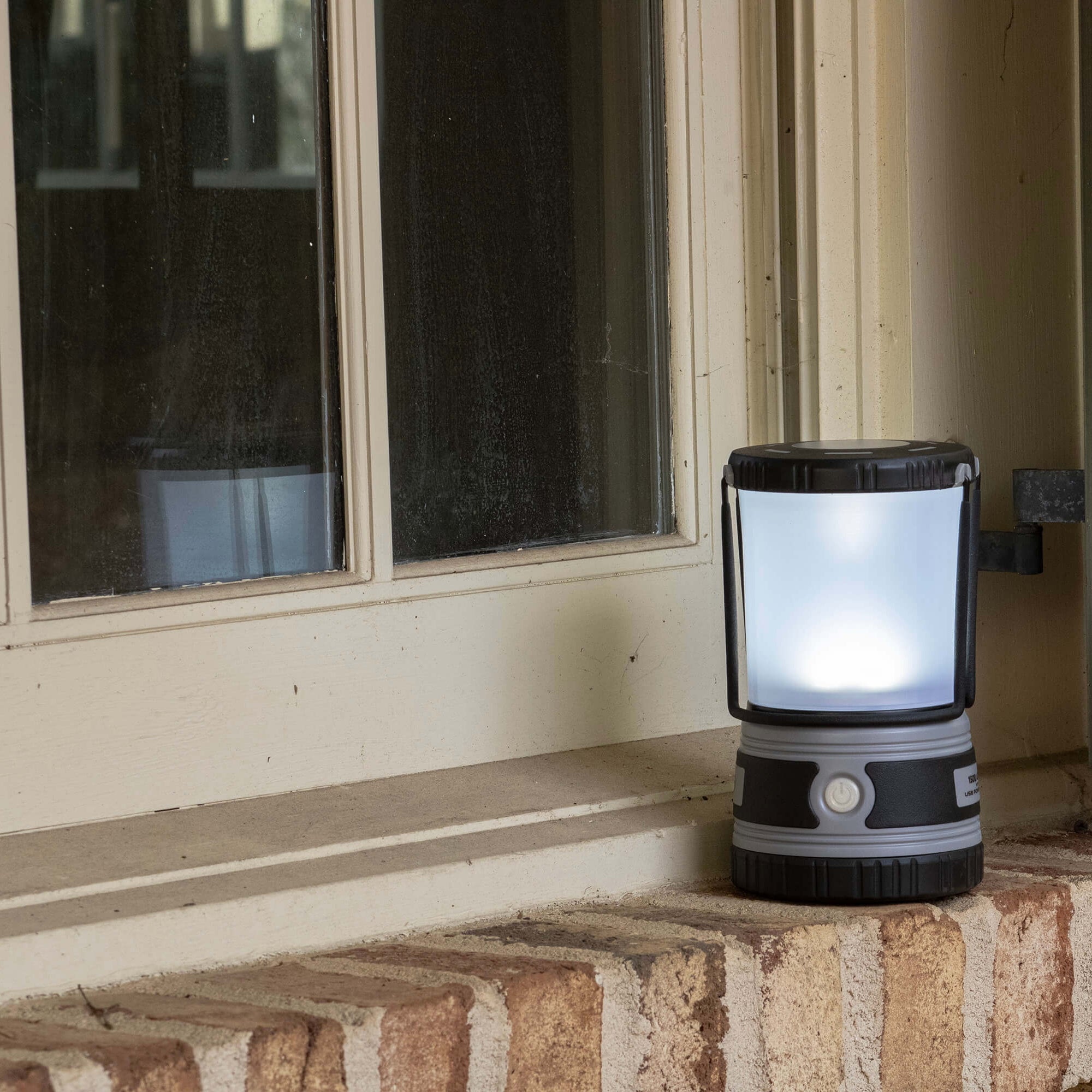 A portable outdoor lantern light with a glowing white LED panel, mounted on a brick ledge next to a window.