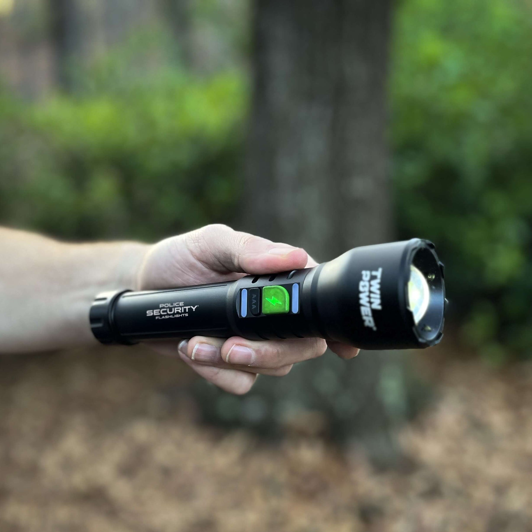 A hand holding a black tactical flashlight with a green button and main LED light illuminated, set against a blurred outdoor background of trees and mulch.