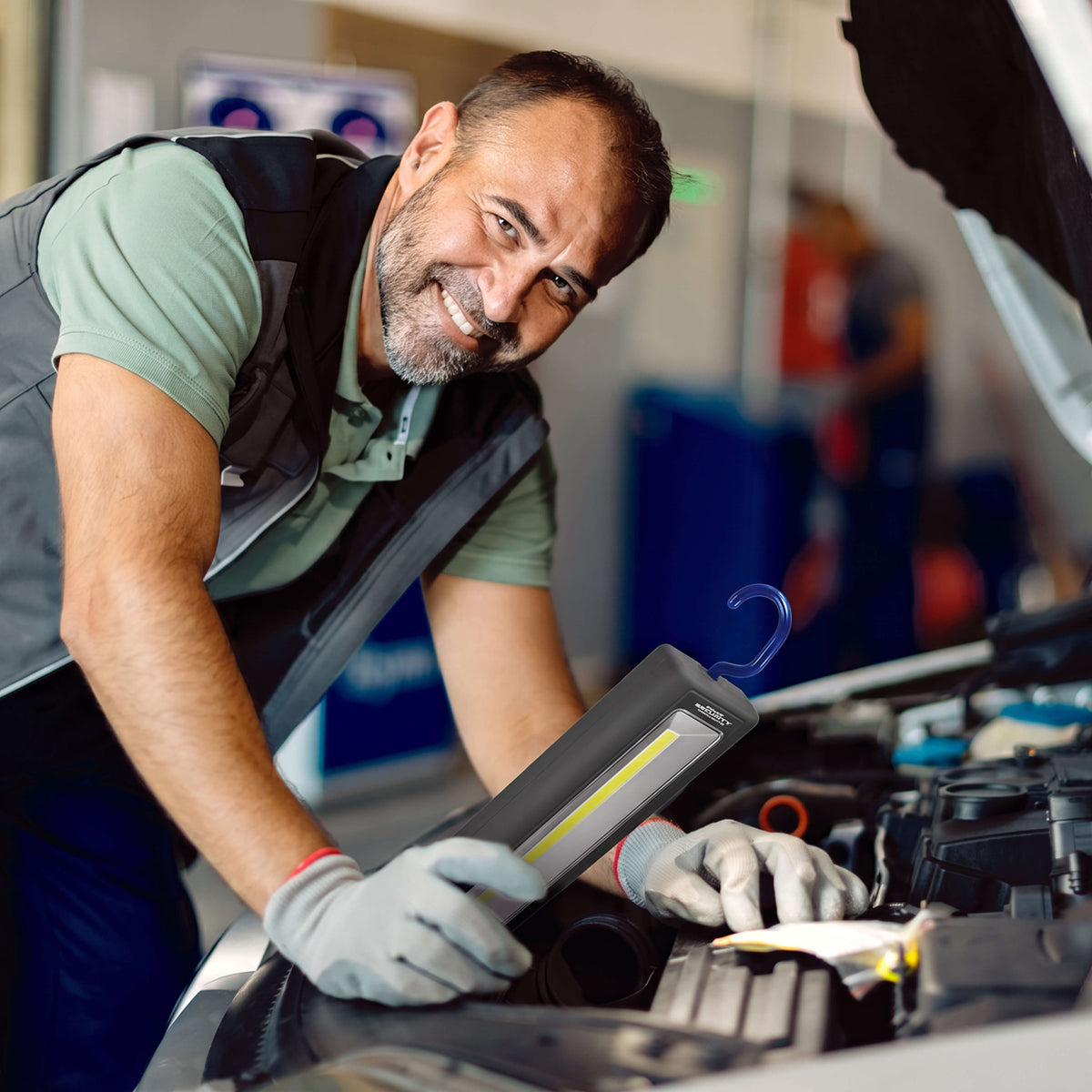 A male mechanic in a garage smiles while inspecting a car engine, holding a Widescope work light. He is wearing a gray utility vest over a green t-shirt and blue pants, with white gloves on, next to an open car hood.