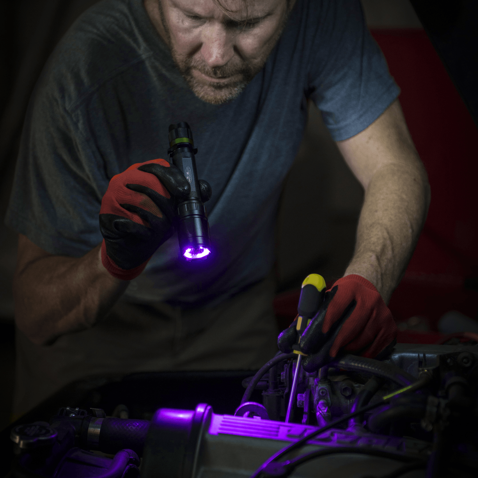 A man in a garage wearing red gloves inspects a car engine, holding a black Police Security Trac Tact-R UV flashlight that casts a bright purple UV glow over the engine components.