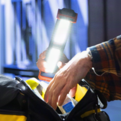 A person's hand holding a bright, illuminated work light next to a black utility bag with yellow accents, set against a blurred blue-toned outdoor background.