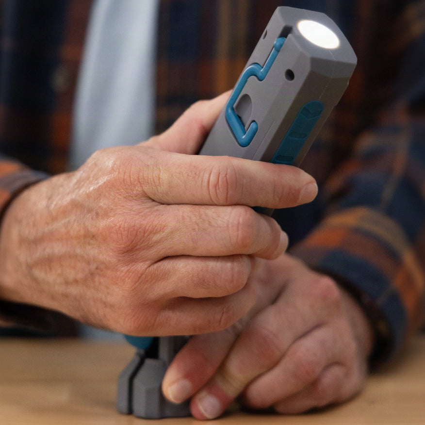 A close-up of a person's hands holding a gray and blue cordless flashlight with its light turned on, wearing a blue and orange plaid flannel shirt.