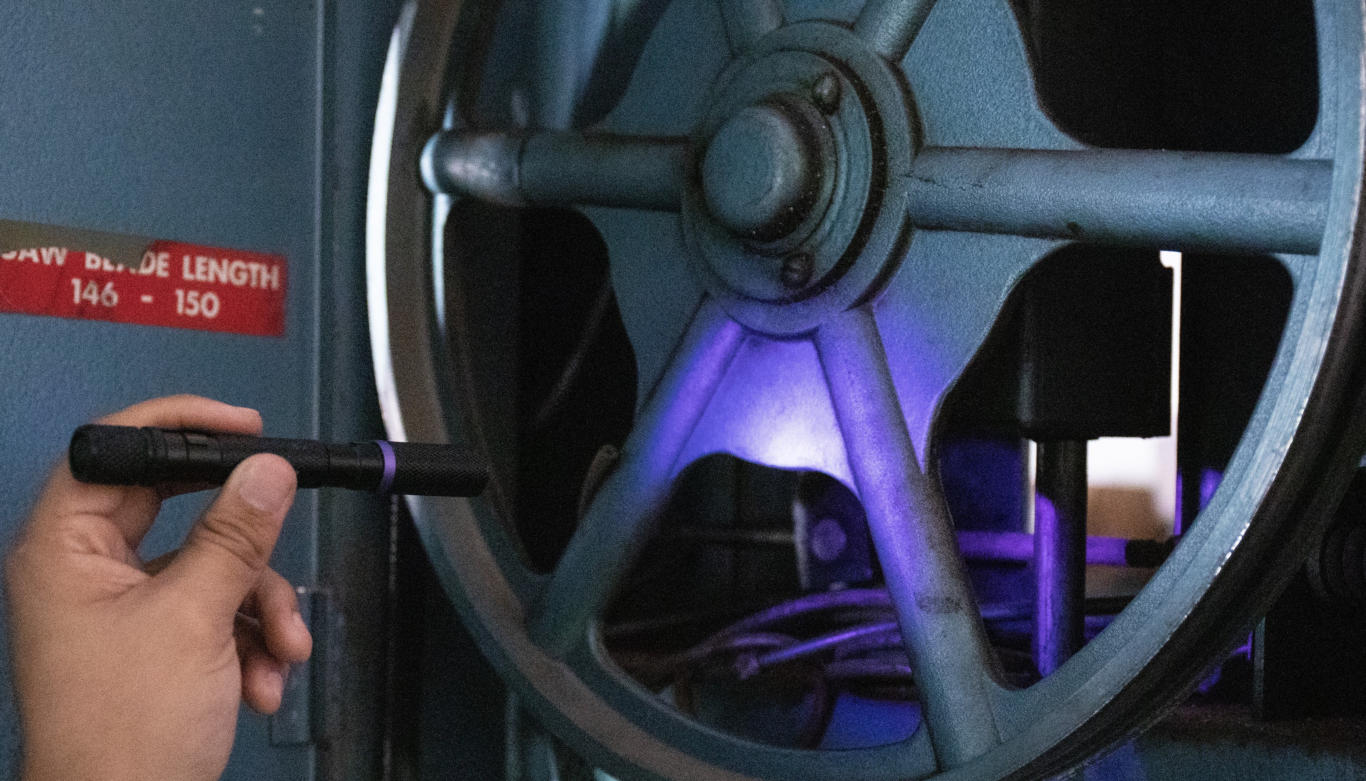 A hand holding a black cylindrical flashlight shining UV light toward a large blue-gray wheel, with a purple glow illuminating the wheel and interior machinery in the background.