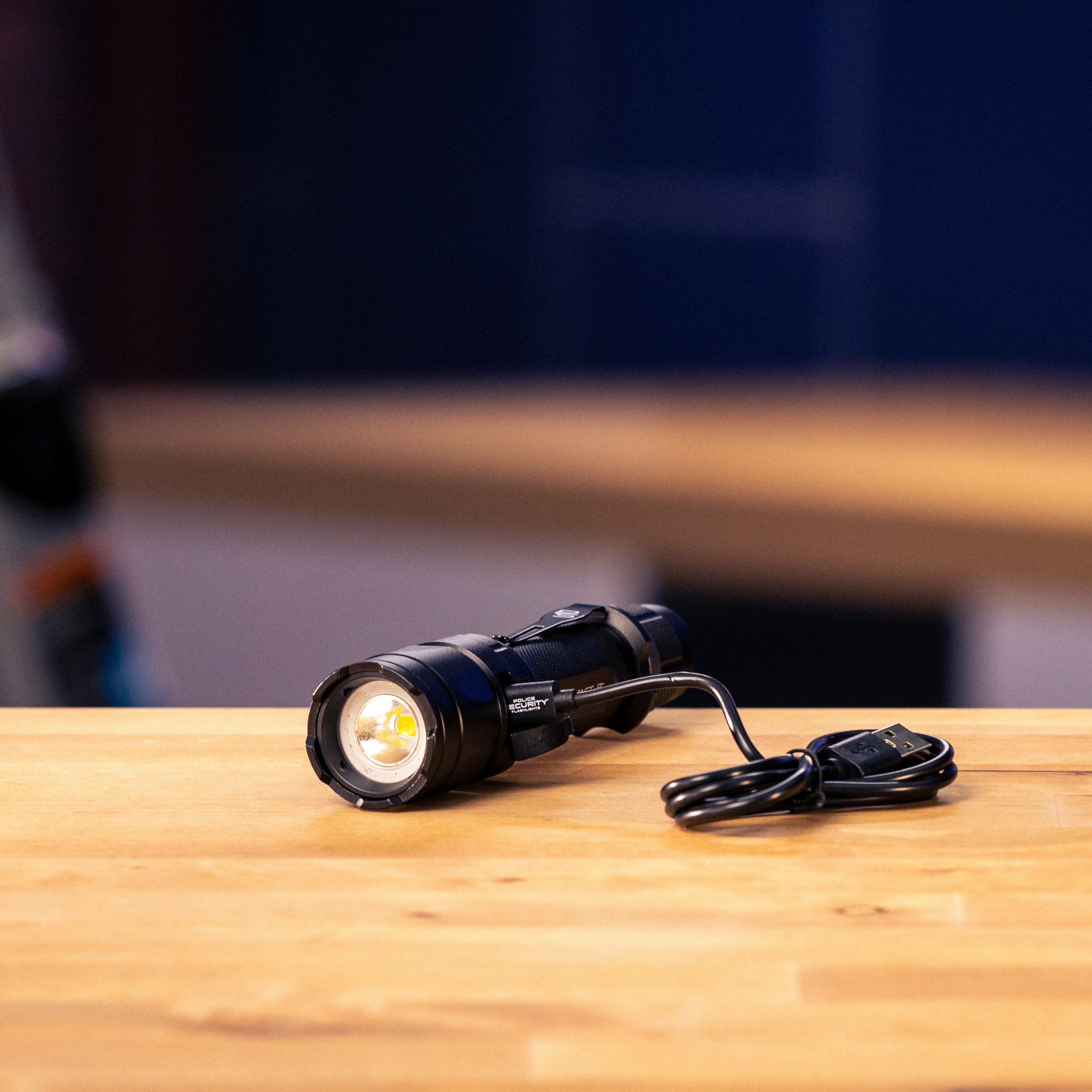 A black Police Security Trac Tact-R tactical LED flashlight with a coiled black power cable, resting on a light-colored wooden tabletop with a blurred dark blue background.