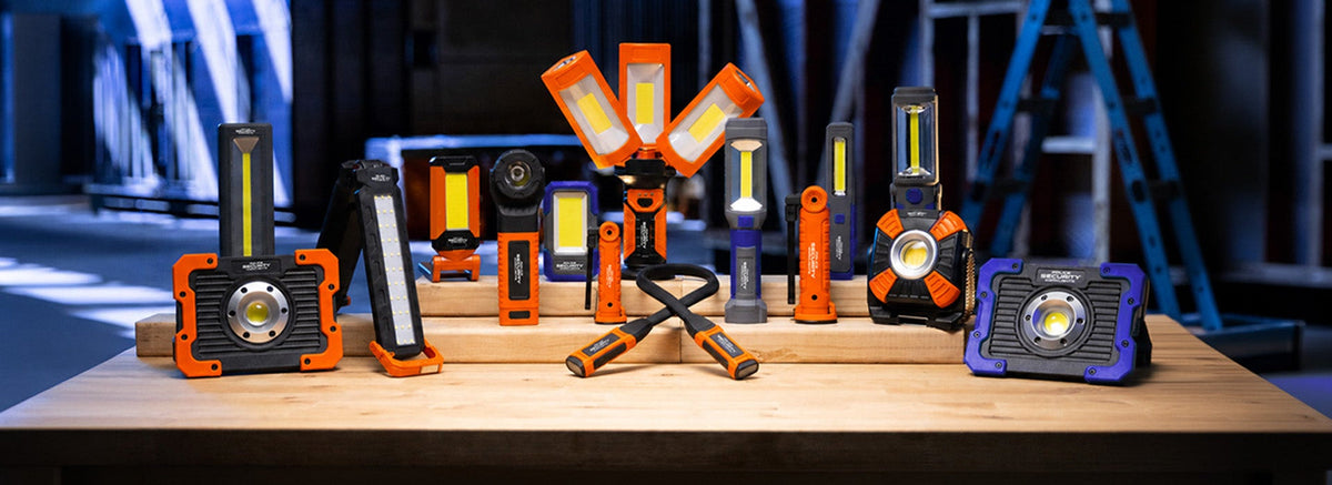 A collection of rugged LED work lights and flashlights in orange, black, gray, and blue, displayed on wooden stands on a workshop table with a blurred industrial background.