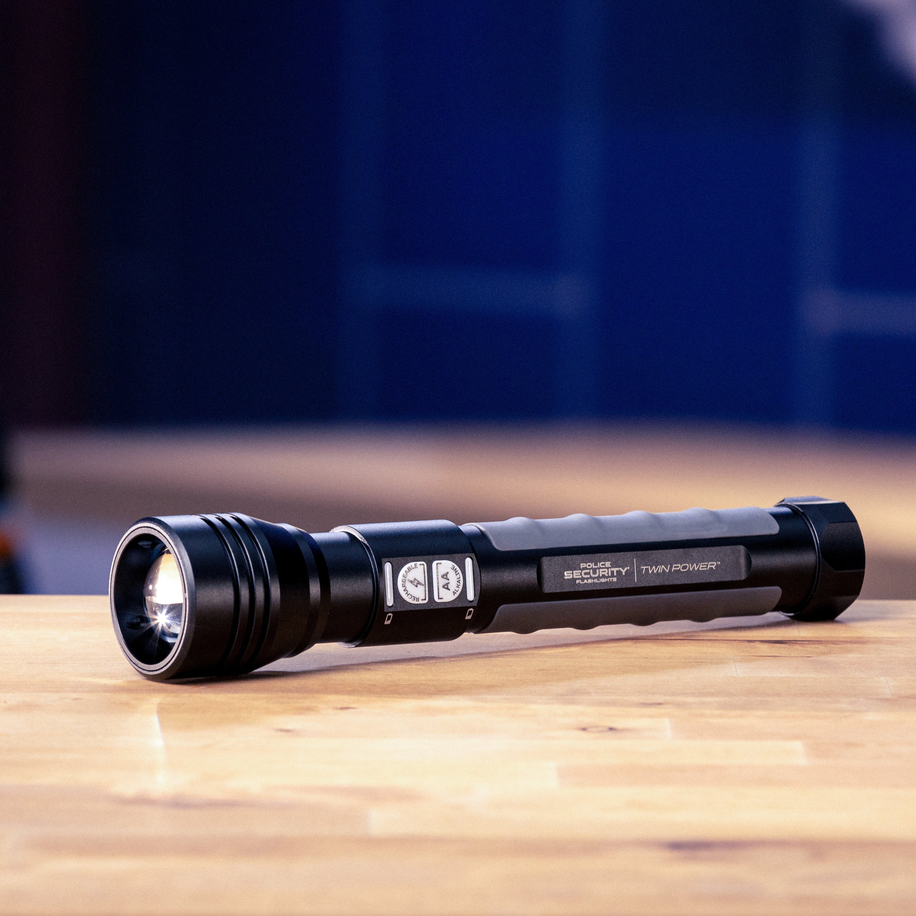 A black Police Security Twin Power flashlight with a textured grip and silver accents, resting on a light wooden tabletop with a blurred workshop background.