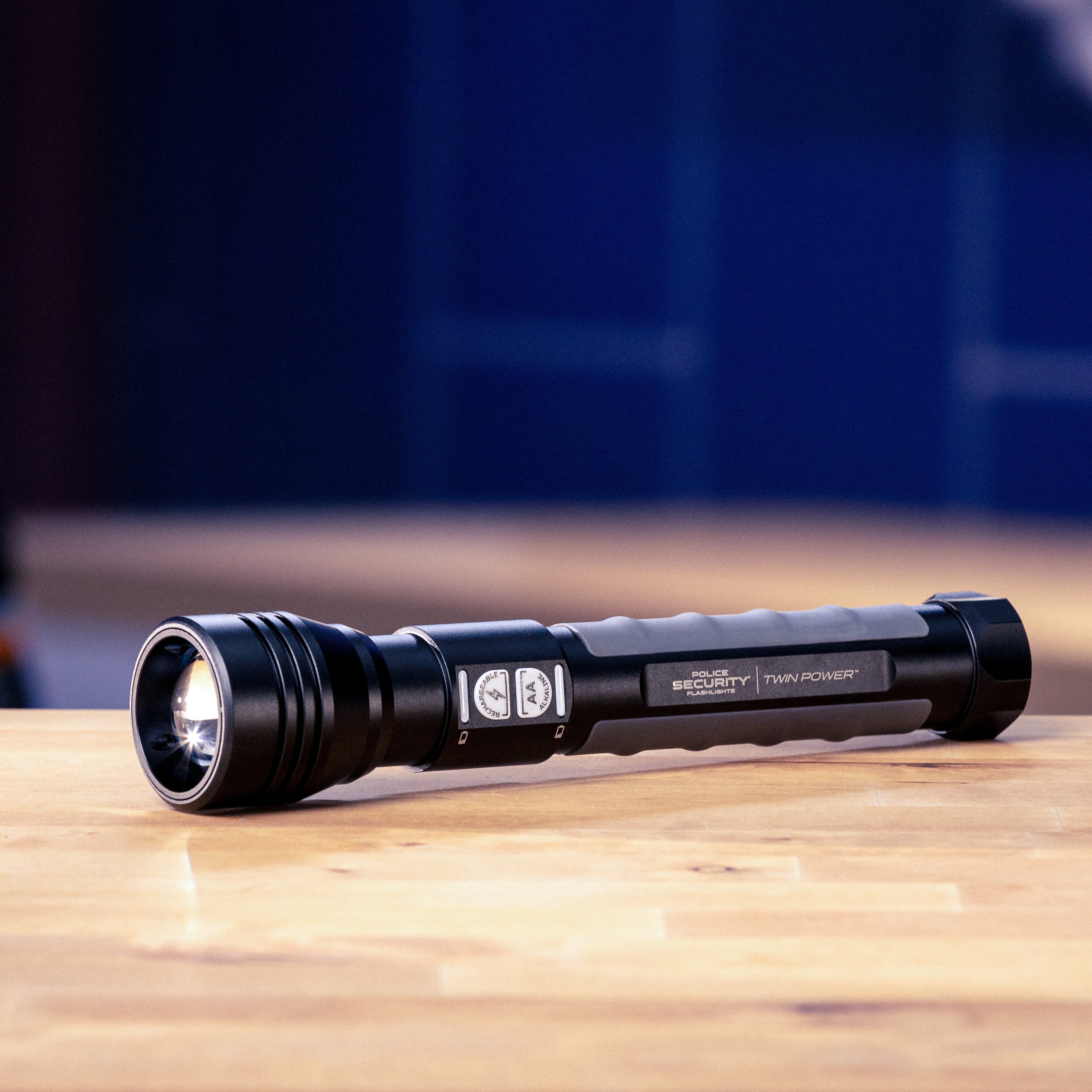 A black Police Security Twin Power flashlight with a textured grip and silver accents, resting on a light wooden tabletop with a blurred workshop background.