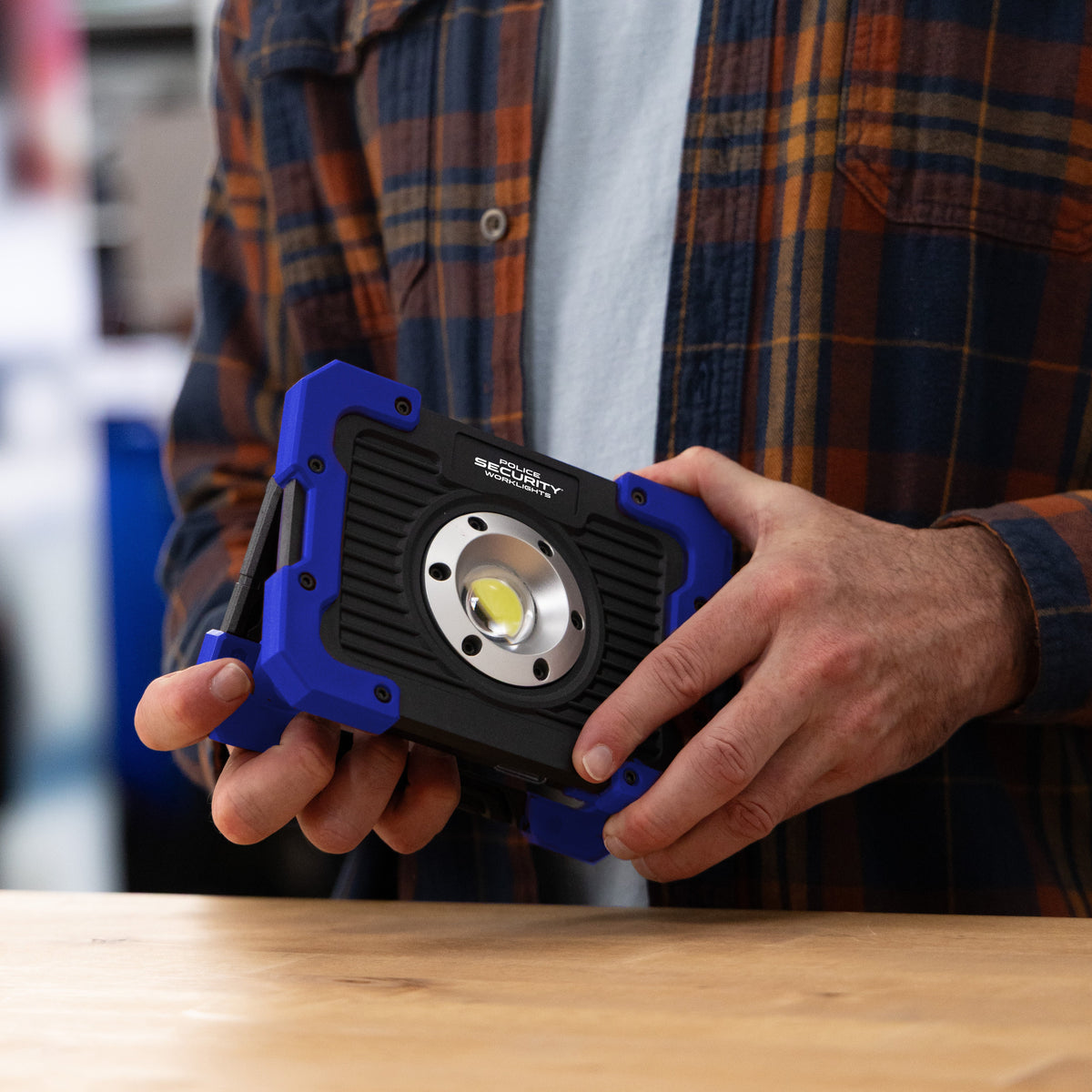 A person in a brown and orange plaid flannel shirt holds a rugged black and blue LED work light with a large circular lens, standing over a wooden table.
