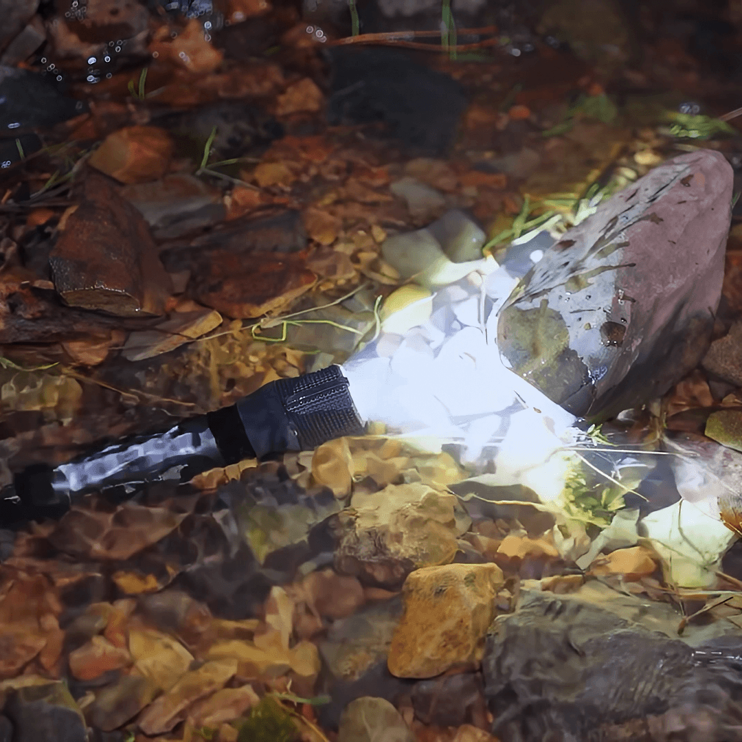 A Police Security Zephyr-R flashlight with a black body, lying in shallow water among rocks and fallen leaves. The light is turned on, casting a bright white glow.