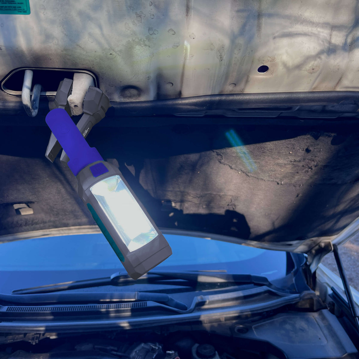 A portable car hood light with a blue handle and a bright white LED lamp, clipped onto the inside of a car hood to illuminate the engine bay.