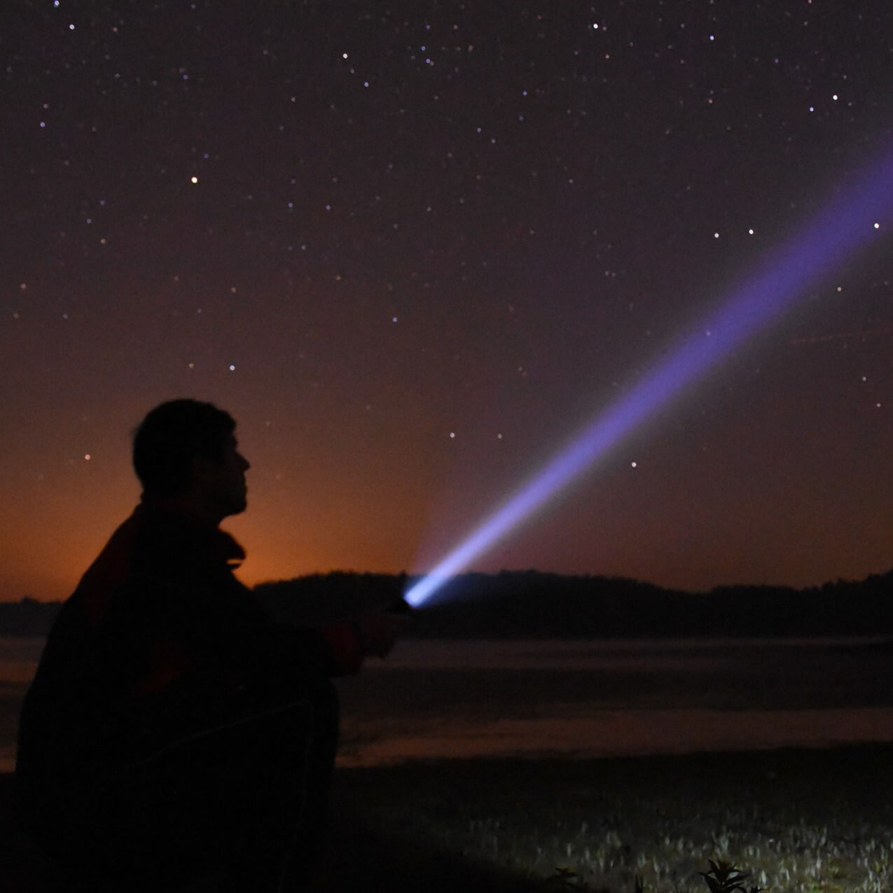 A person in silhouette at night points a powerful beam flashlight or laser toward the stars in a starry sky, with a dark landscape and orange horizon in the background.