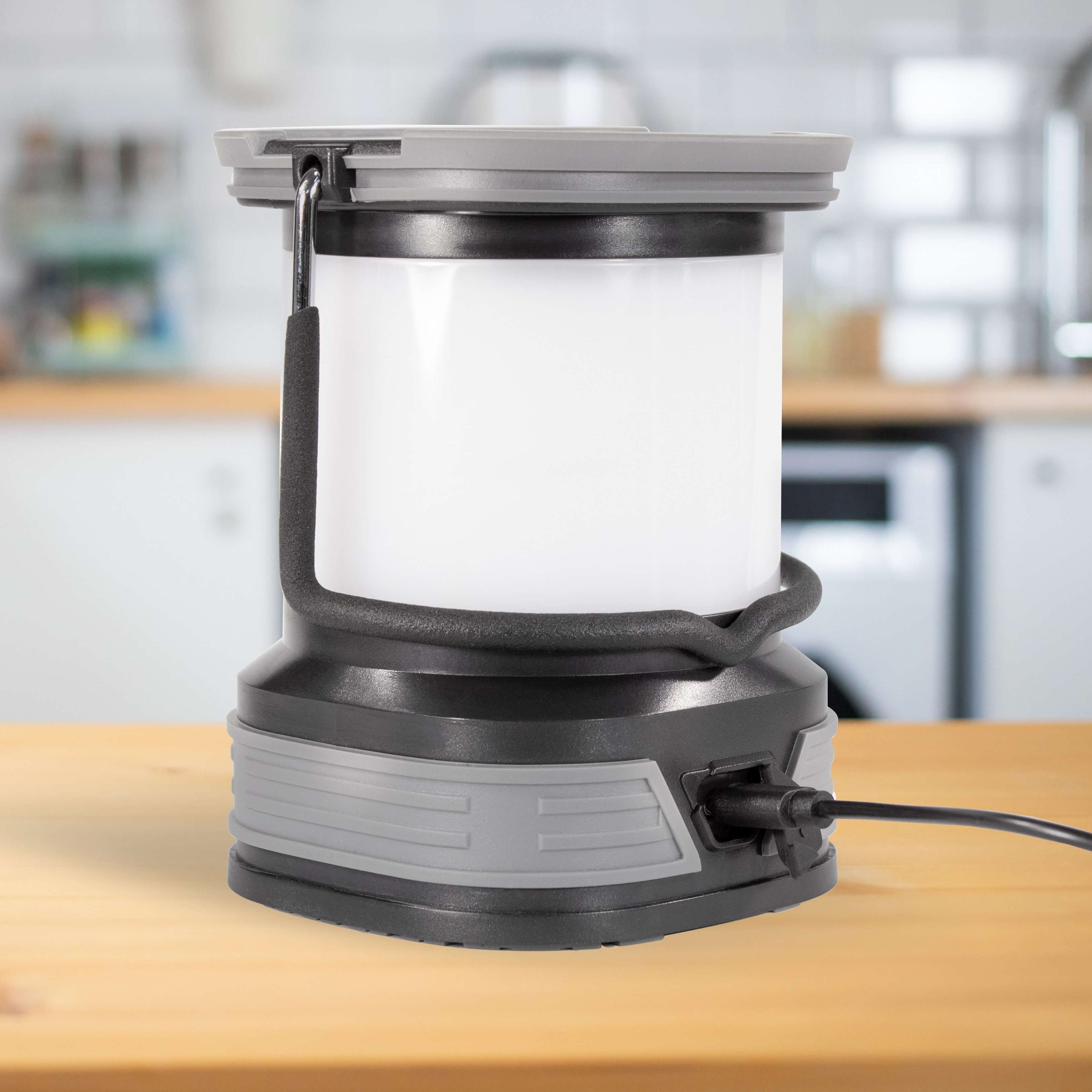 A portable camping lantern with a white translucent body, gray and black base, and carrying handle, shown [lugged in to charge on a wooden countertop with a blurred kitchen background.