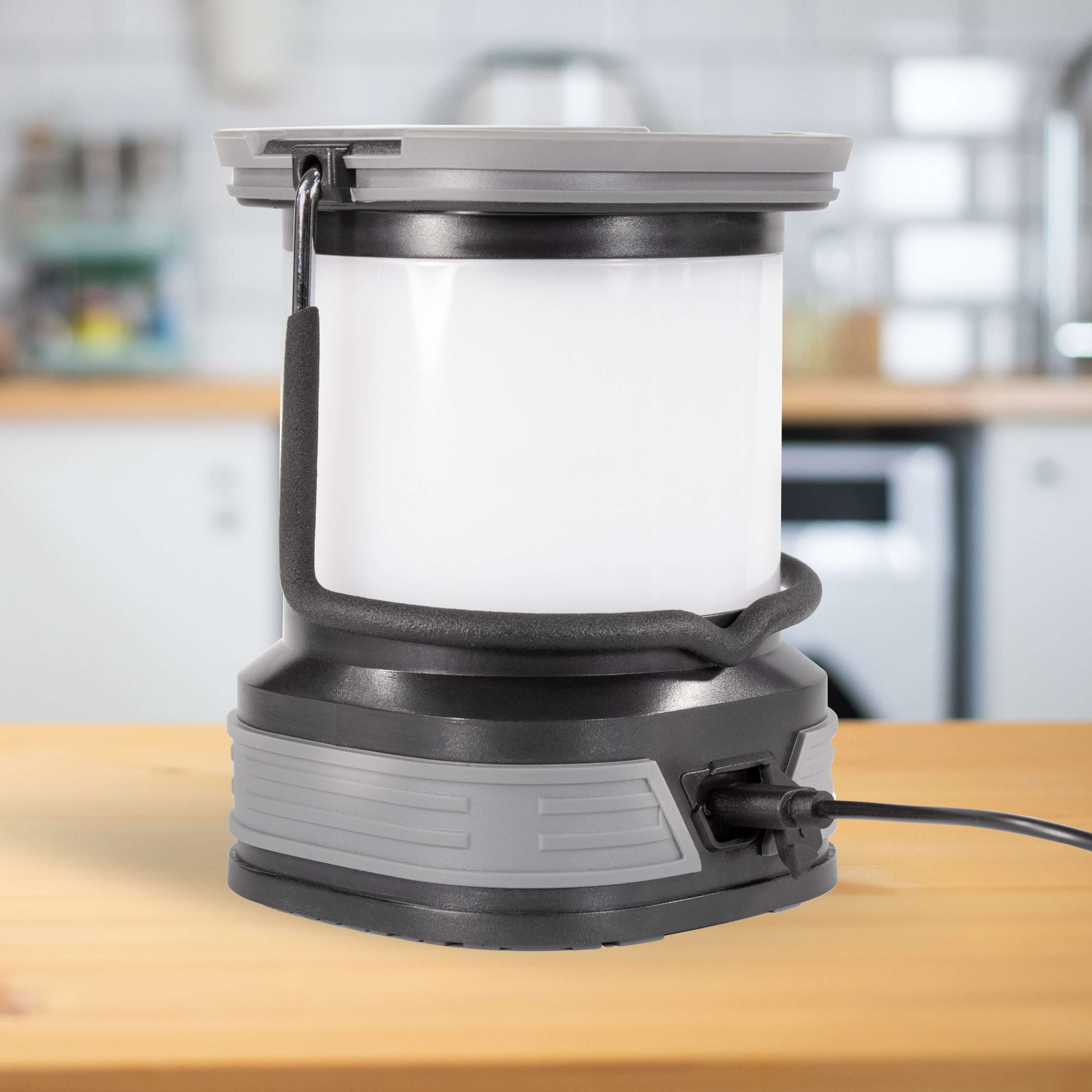 A portable camping lantern with a white translucent body, gray and black base, and carrying handle, shown [lugged in to charge on a wooden countertop with a blurred kitchen background.
