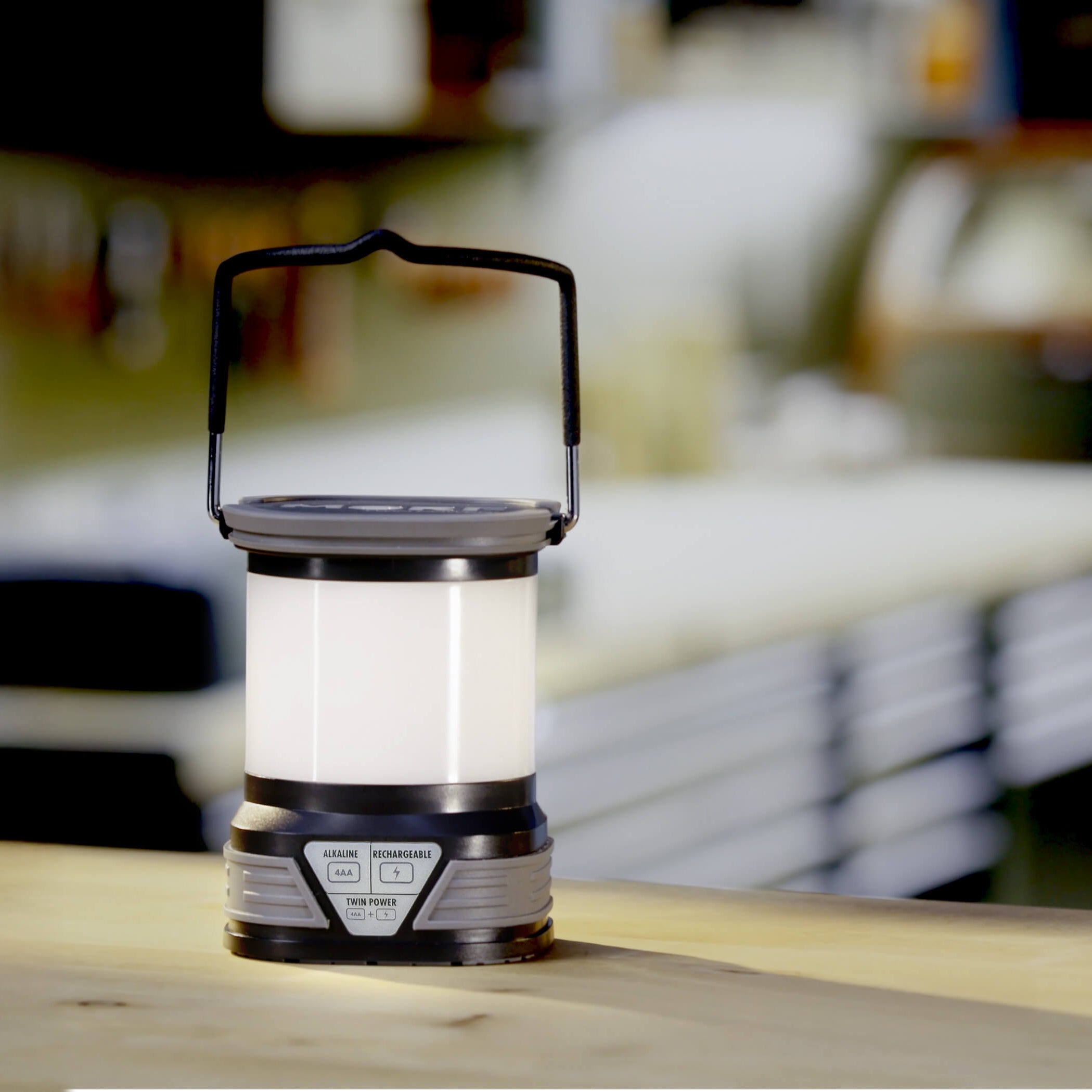 A portable camping lantern with a black handle and silver and black body, standing on a wooden surface with a blurred workshop or garage background.
