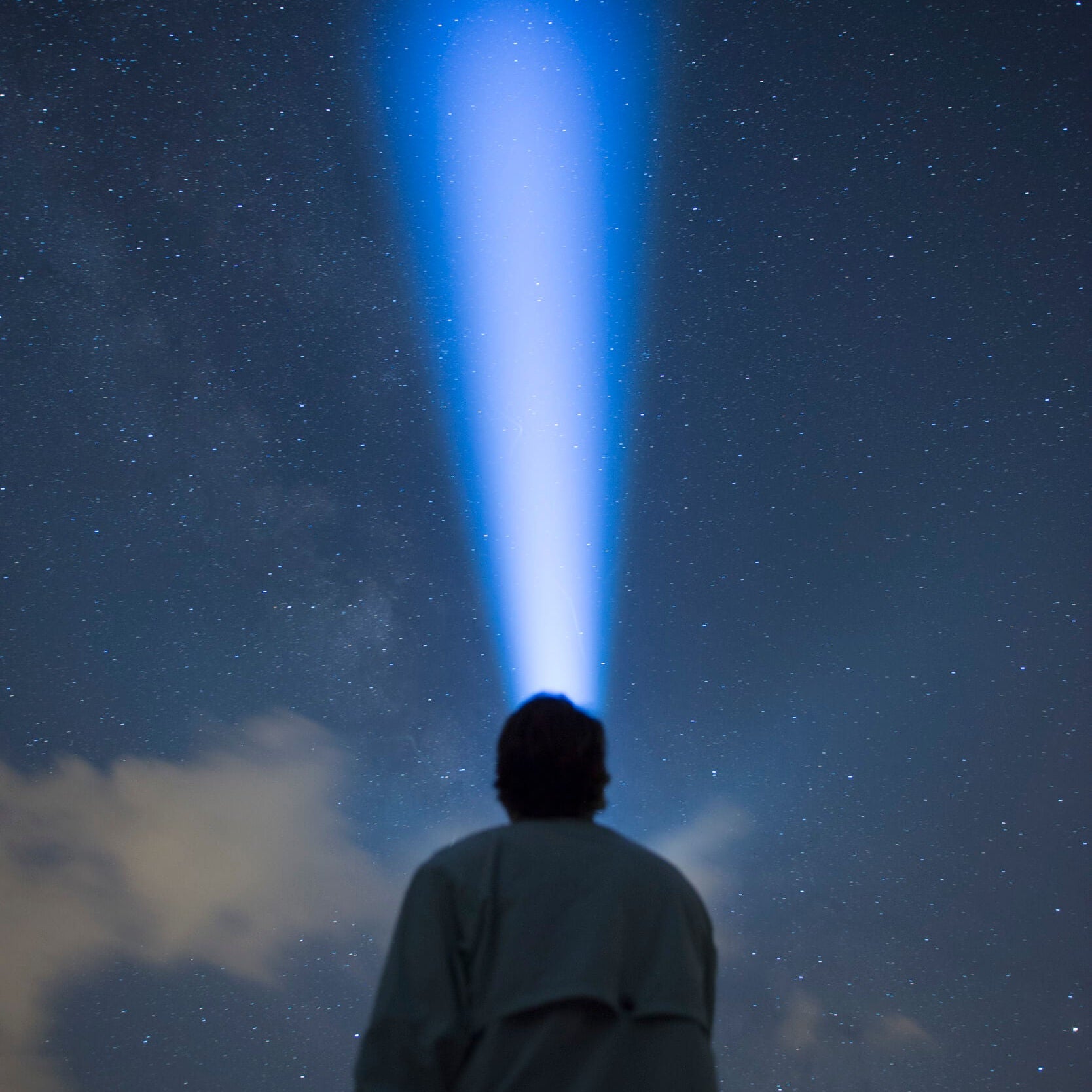 A person seen from behind wearing a light-colored coat, looking up at a starry night sky with a bright blue beam of light shining down toward them.