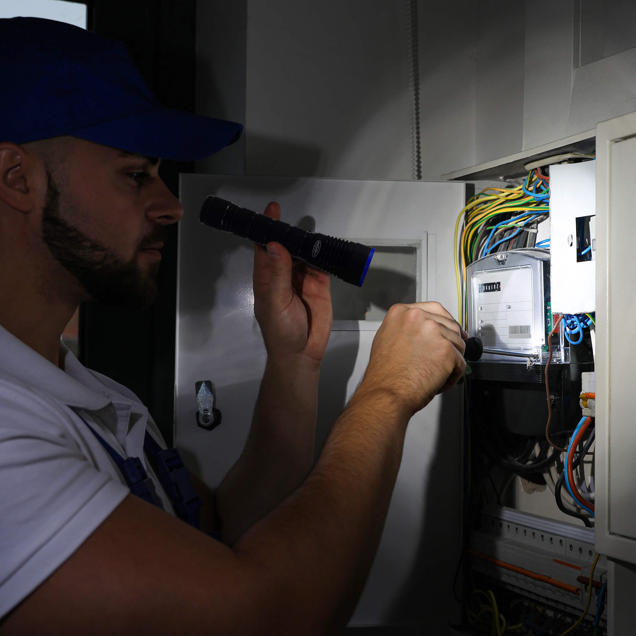 A man in a blue cap and white shirt inspects an electrical panel, holding a black flashlight inside an open cabinet filled with colorful wires and a meter.