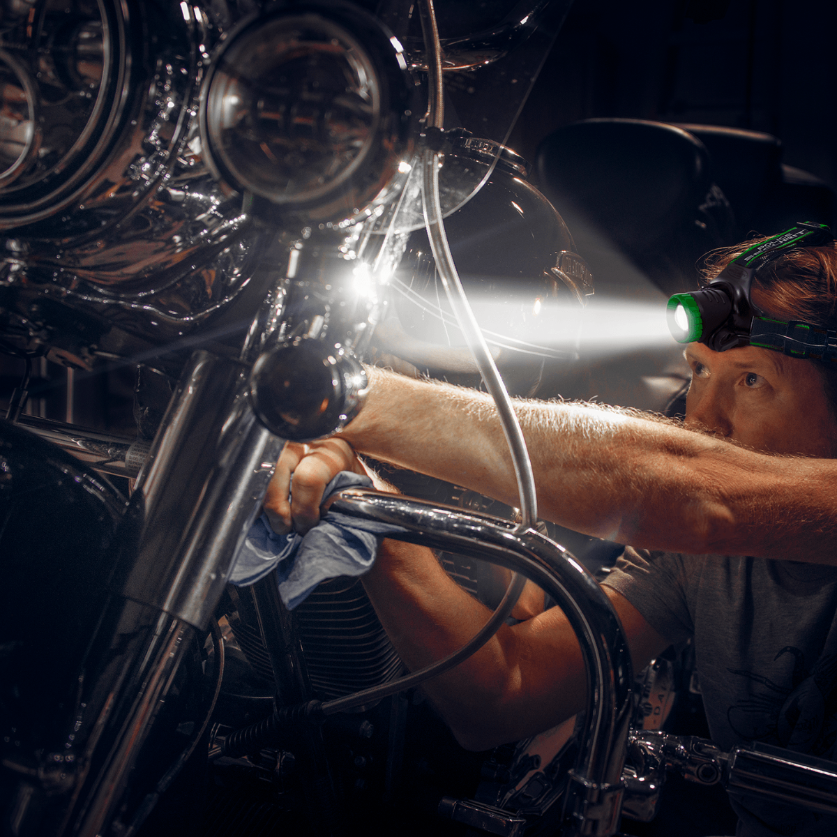 A man wearing a headlamp with a bright green light and a beam of white light works on a motorcycle in a dark setting. The motorcycle has chrome details and dual round headlights.