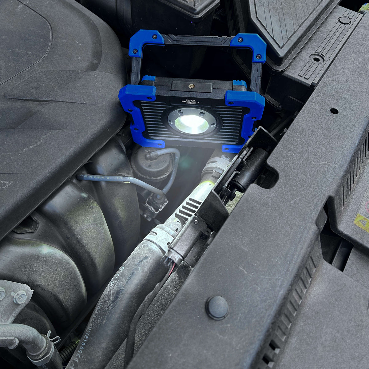 A rugged blue and black portable work light with a bright illuminated LED, clipped onto a car engine bay.