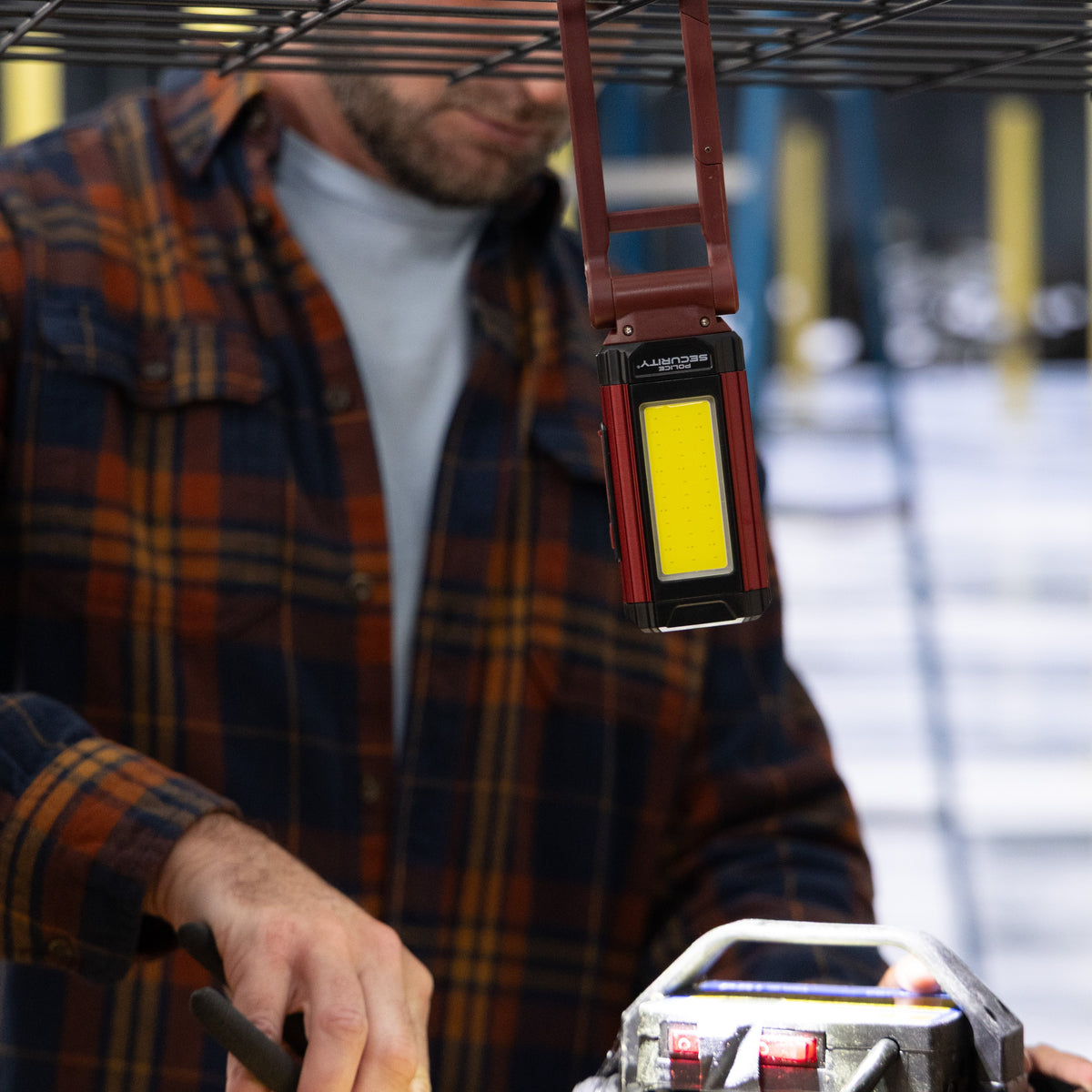 A man in a brown and orange plaid flannel shirt works under a black metal wire rack, holding a pair of pliers near a workbench with a power tool. A red and black cordless lantern hangs from the wire rack, illuminating the workspace.
