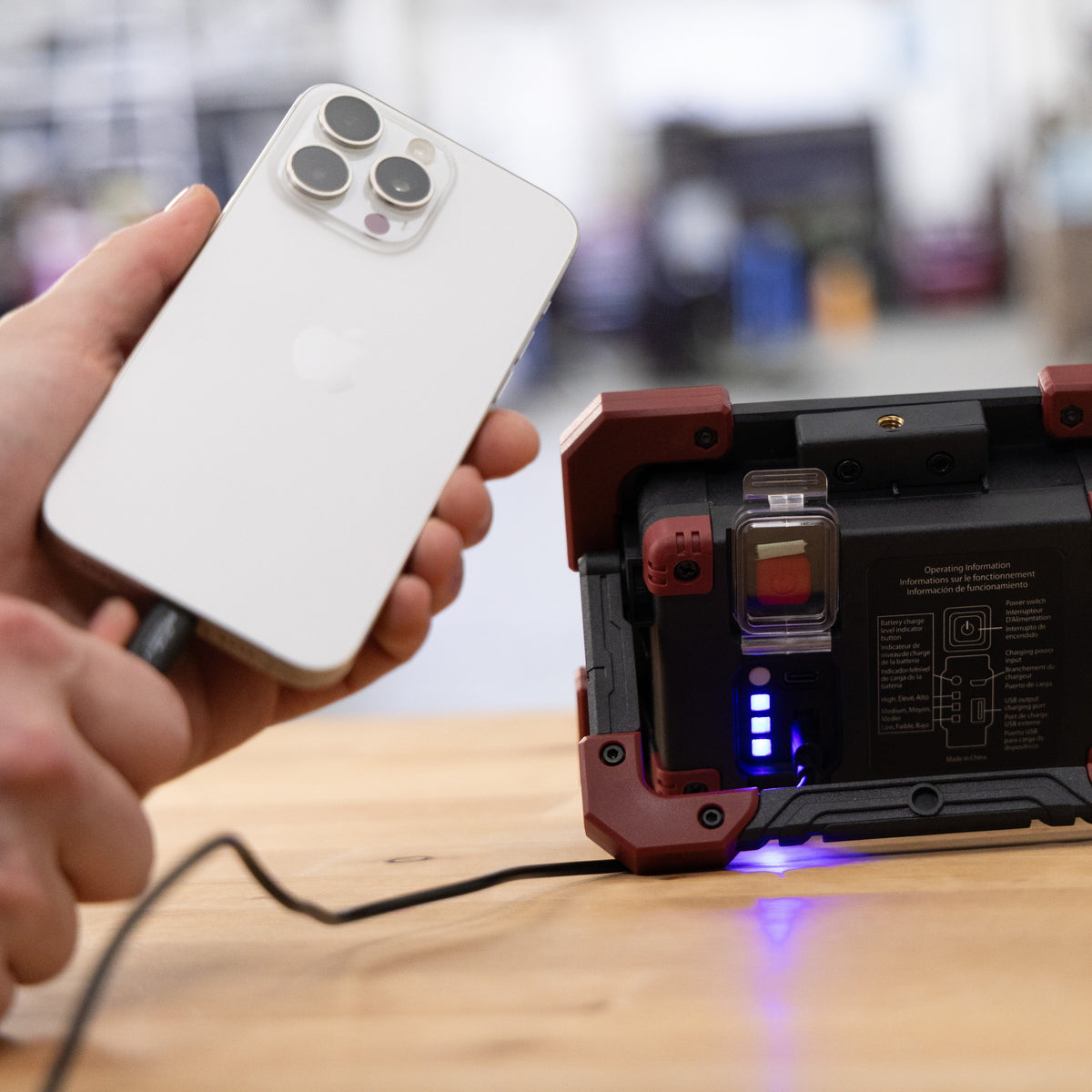 A hand holds a white smartphone being charged from the red and black Kickstand-R Worklight on a wooden table. The device has a black interior with blue LED lights and a transparent component, set against a blurred indoor background.