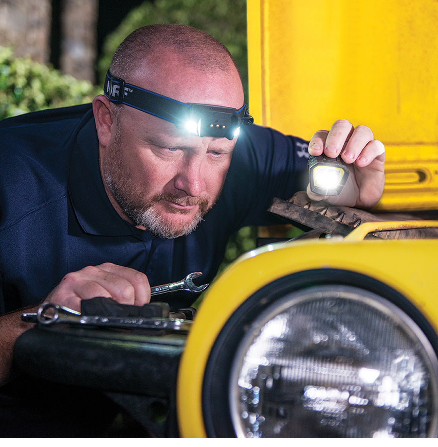 A man wearing a headlamp and using the lamp from its headlamp to see the parts of the car he's fixing