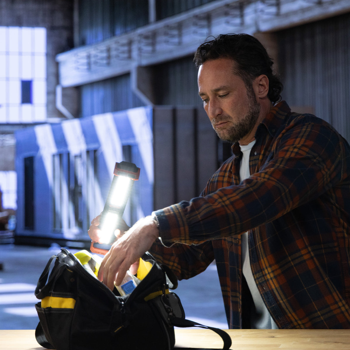 A man in a brown and orange plaid flannel shirt reaches into a black tool bag with yellow accents on a wooden workbench inside a workshop.