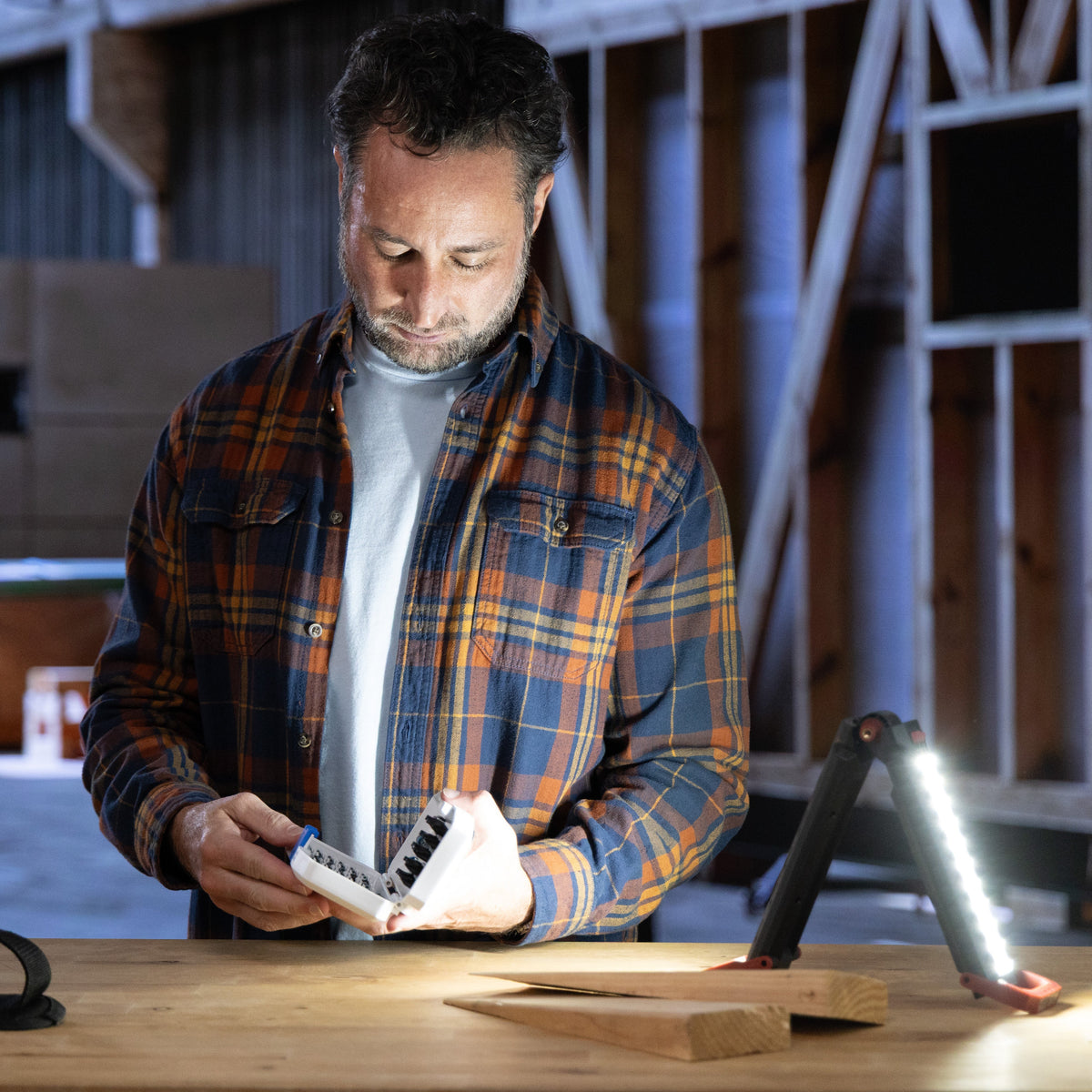 A man in a workshop wearing a blue and orange plaid flannel shirt, holding a set of drill bits and looking down at it. A black tool bag sits on the table to his left, and a bright work light is turned on to his right.