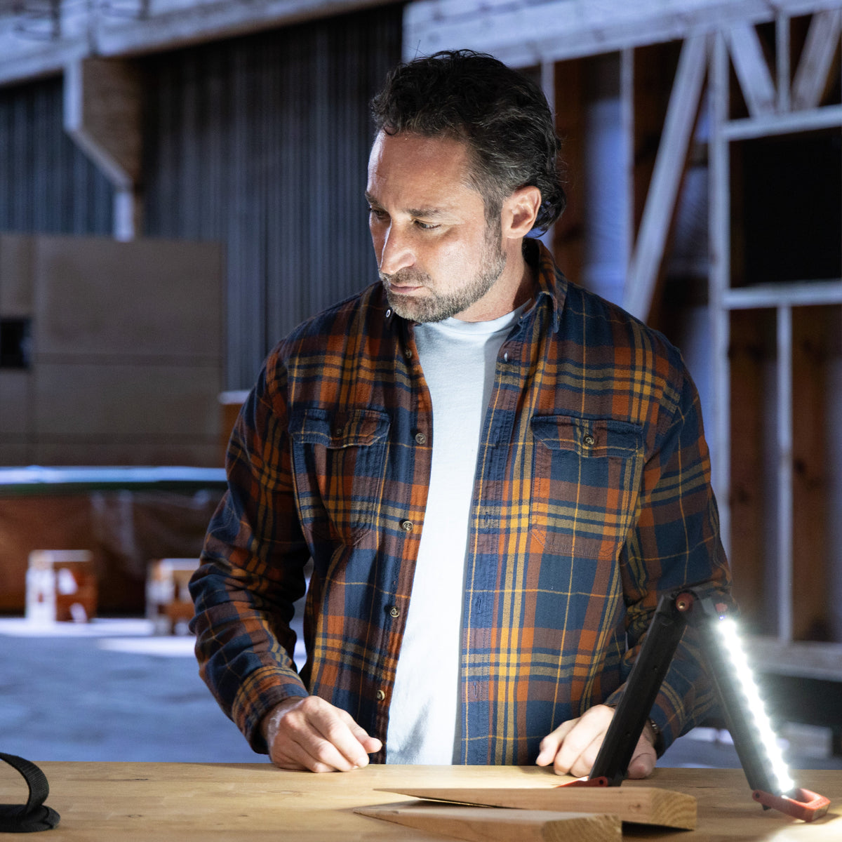 A man in a blue and orange plaid flannel shirt stands at a workbench in a workshop, with holding a bright LED work light standing on a wooden table. A black and yellow tool bag is visible on the left.