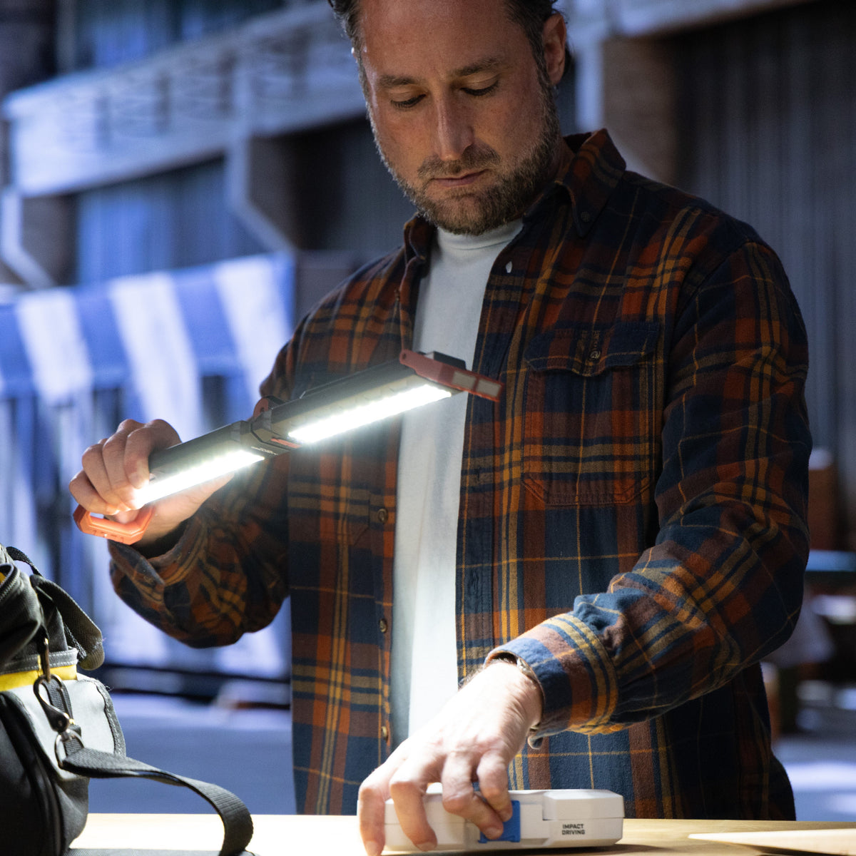 A man in a brown and orange plaid flannel shirt uses a bright LED flashlight to illuminate a folding work light on a workbench in an industrial workshop setting.