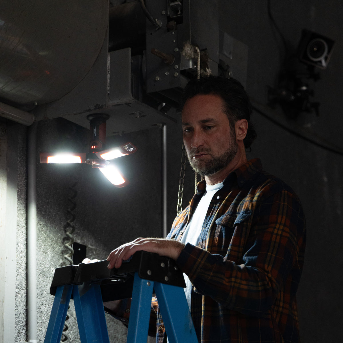 A man in a brown and black plaid flannel shirt stands on a blue step ladder in a dark industrial room, looking down at the ladder. A bright work light is turned on above him, casting strong shadows on the concrete wall behind him.